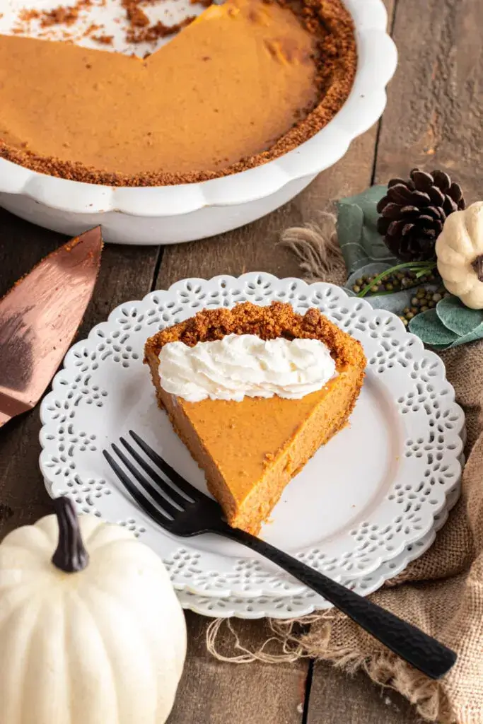 Pumpkin pie with graham cracker crust on a white plate.  A dollop of whipped cream sits on top of the wedge of pie and a fork rests next to it.