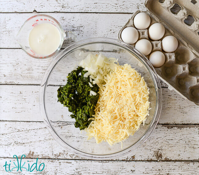 Quiche ingredients, spinach, cheese, onions, cream, and eggs, arranged on a weathered white wooden background
