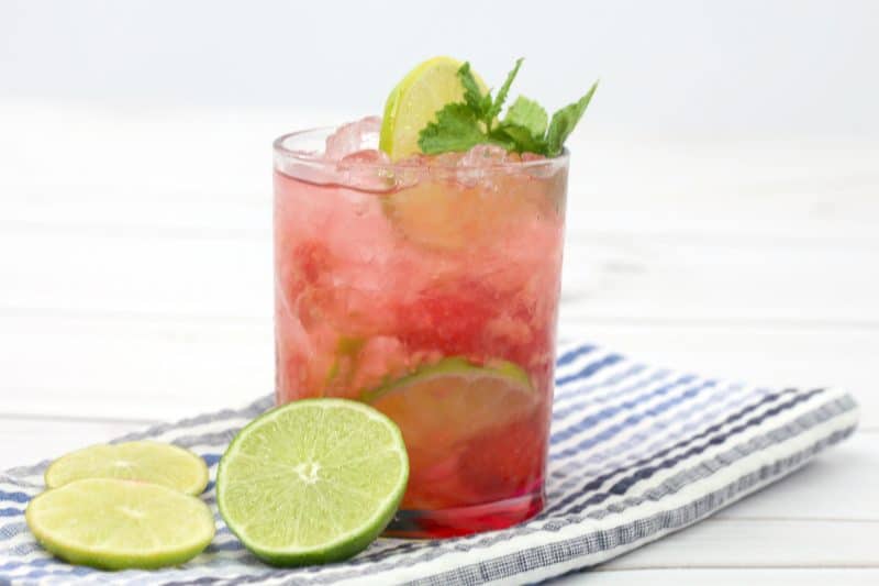 A dark pink raspberry lime ricky cocktail in a lowball glass filled with crushed ice, and garnished with a slice of lime and fresh mint.  The glass sits on a grey and white striped tea towel, and sits next to three slices of lime.