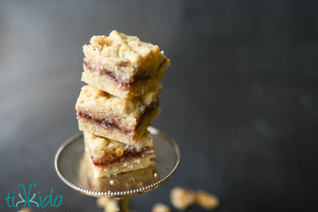 Raspberry crumble bar cookies stacked on a tiny cake stand, surrounded by cookies and walnuts on a black chalkboard background.