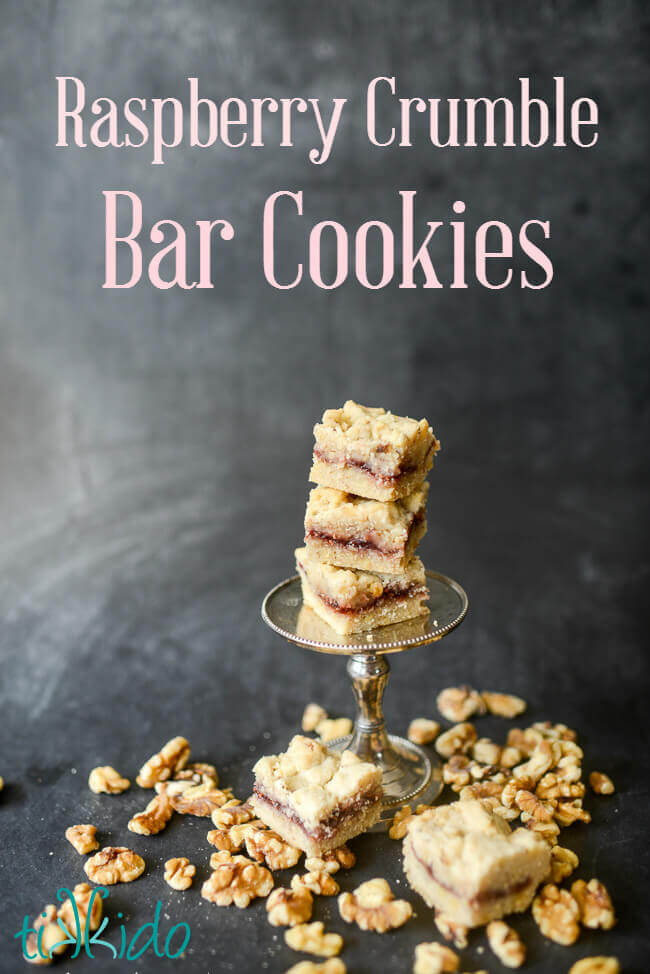 Raspberry crumble bar cookies stacked on a tiny cake stand, surrounded by cookies and walnuts on a black chalkboard background.