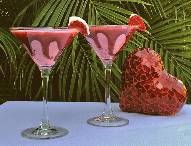 Two red velvet martinis garnished with small slices of red velvet cake, on a white tablecloth.  A red mosaic heart sits on the table, and the background is made from palm fronds.
