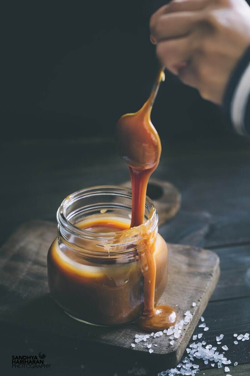 Salted caramel ice cream sauce being spooned into a glass jar, with large sea salt crystals scattered on a cutting board next to the jar.