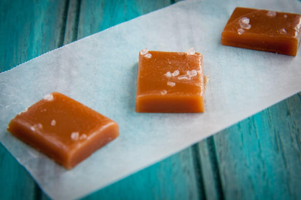 Three honey caramels topped with sea salt crystals on a piece of parchment paper on a blue wooden surface.