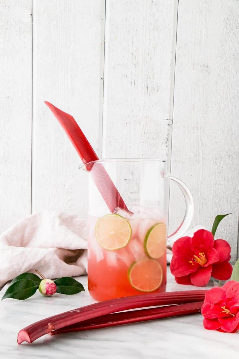 Pitcher of pink rhubarb margarita with ice and slices of fresh lime, and with a stick of fresh rhubarb sticking out of the pitcher.  Fresh flowers and more fresh rhubarb stalks surround the pitcher, which sits on a white marble surface.