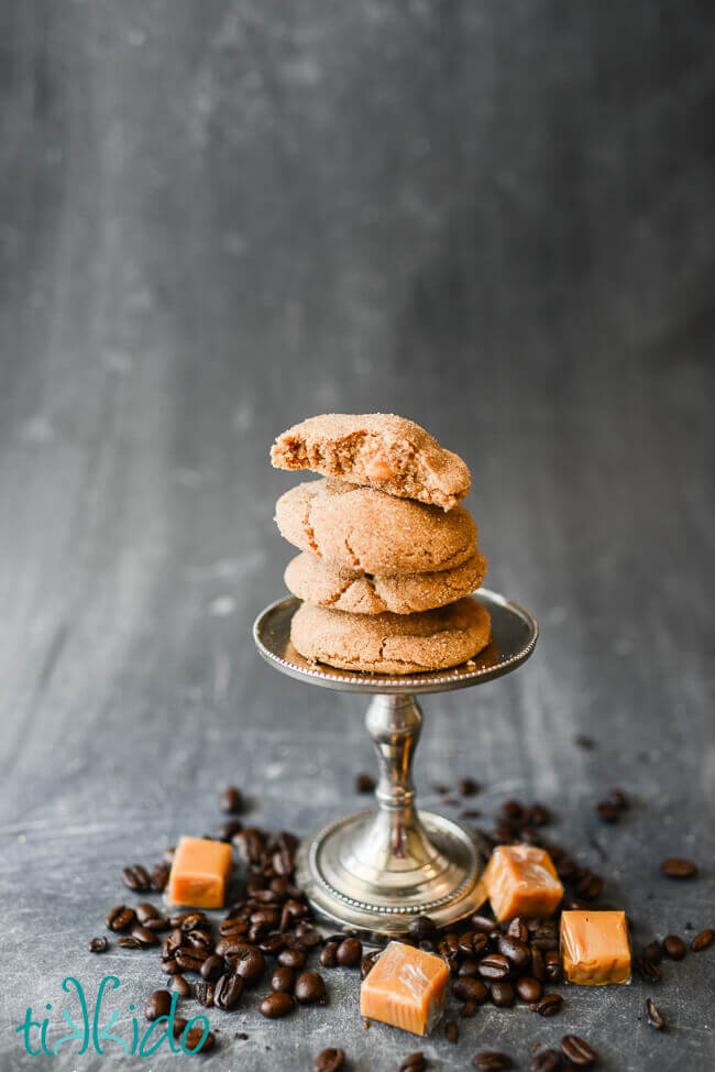 Coffee flavored cookies filled with caramel stacked on a small stand, surrounded by coffee beans and caramel candies.