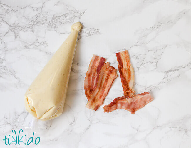 Pancake batter in a disposable piping bag next to strips of cooked bacon.