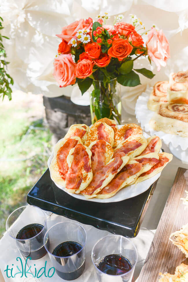 Bacon pancakes on a white plate next to clear glasses with real maple syrup for dipping.