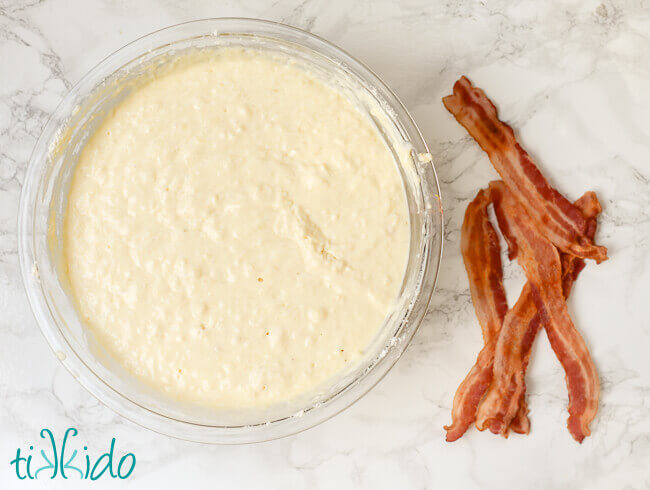 Pancake batter in a clear glass bowl next to strips of cooked bacon.