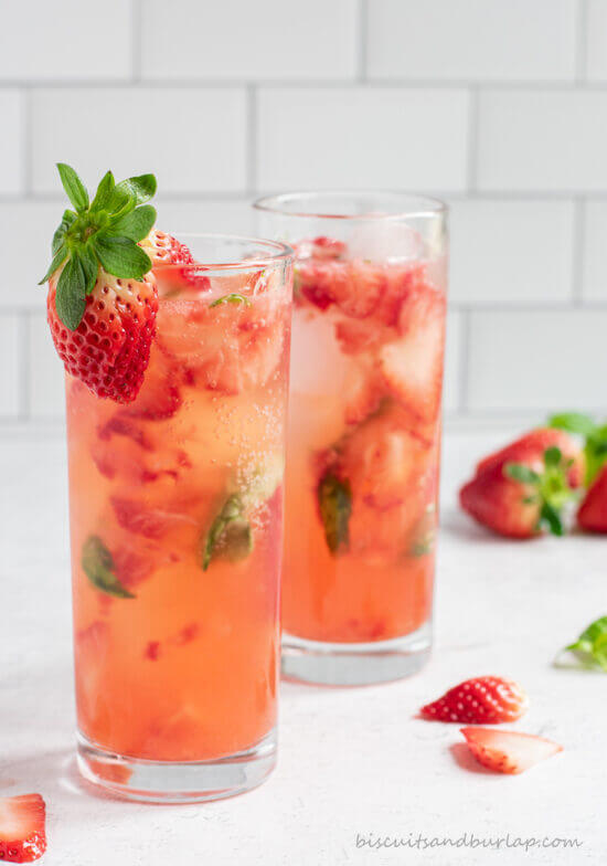 Two glasses of deep pink strawberry basil gin cocktail, garnished with a fresh strawberry, on a white surface.