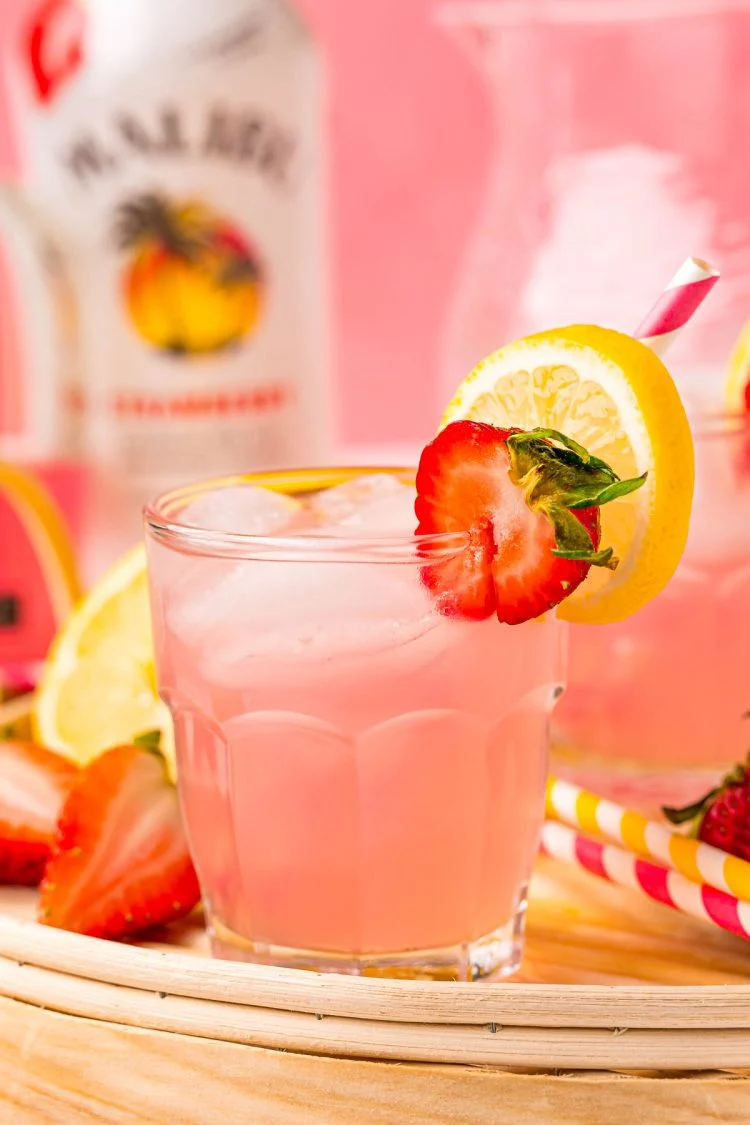 Pink lemonade vodka cocktail in a glass full of ice, garnished with a sliced strawberry, and slice of lemon, and a pink and white paper straw.