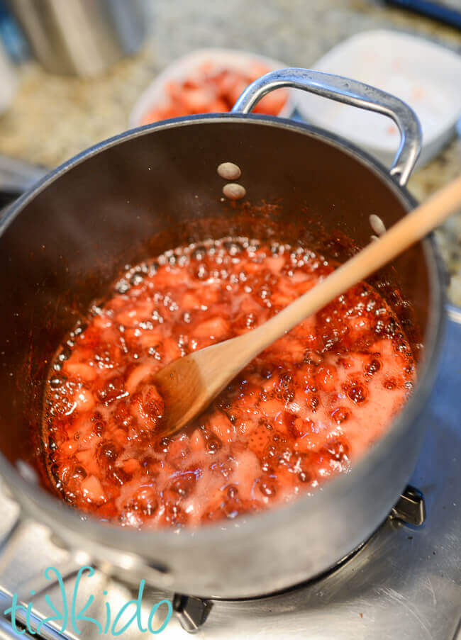 Homemade strawberry sauce being cooked in a pot on the stovetop, with a wooden spoon in the pot.