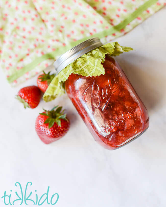 Homemade strawberry sauce in a mason jar, next to strawberries and a strawberry print apron.