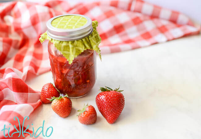 Homemade Strawberry Sauce in a mason jar on a white surface covered with red checkered tablecloth and fresh strawberries.