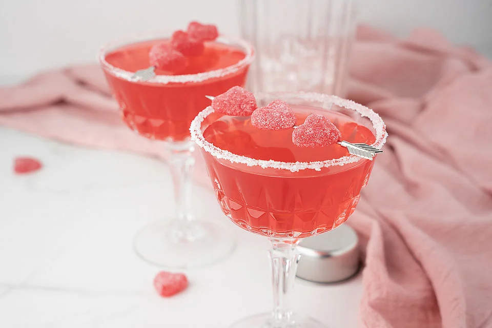Two glasses of deep pink Strawberry Sour Candy Cocktail garnished with a silver cocktail arrow spearing three heart shaped candies.