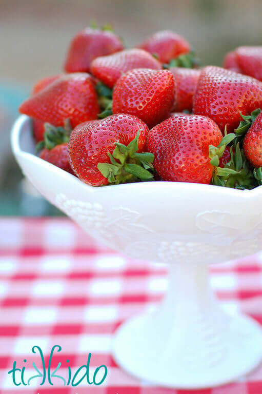 White milk glass compote filled with fresh strawberries on a red and white checkered tablecloth.