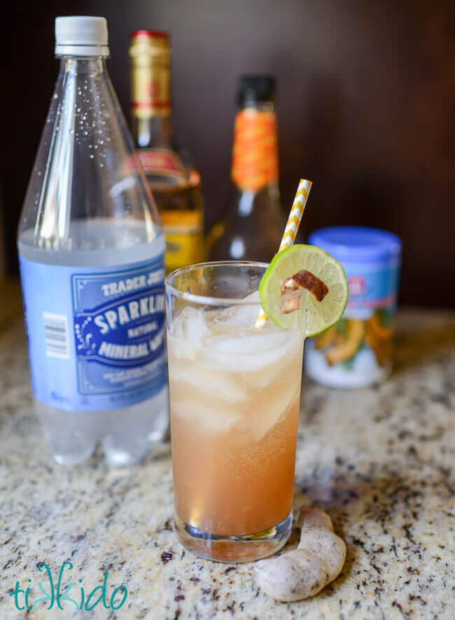 Tamarind margarita in a tall glass with a yellow and white striped paper straw, slice of lime garnish, and peel of tamarind garnish.  Limes and tamarind pods on wooden surface around glass.