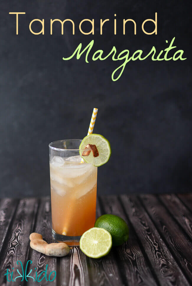 Tamarind margarita in a tall glass with a yellow and white striped paper straw, slice of lime garnish, and peel of tamarind garnish.  Limes and tamarind pods on wooden surface around glass.