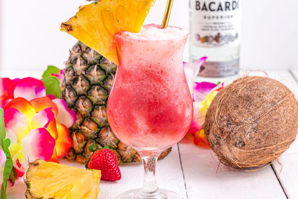 Pink and white Lava Flow Cocktail in a hurricane glass, garnished with a wedge of pineapple.  A fresh pineapple, whole coconut, and tropical flowers are in the background.