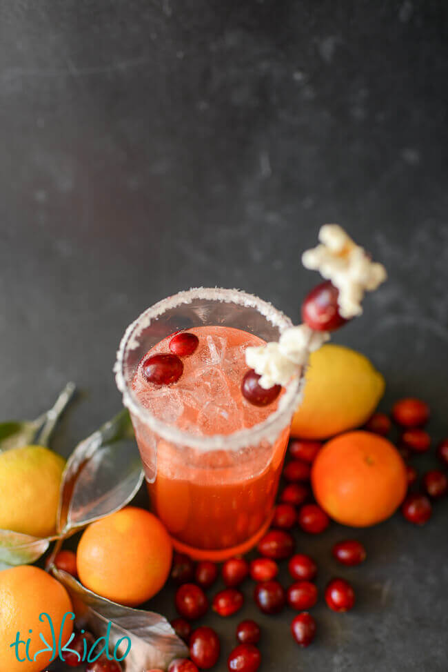 Merry and Bright Citrus Christmas Cocktail garnished with fresh cranberries and popcorn on a stick, on a black chalkboard background.  Fresh oranges, lemons, and cranberries surround the glass.