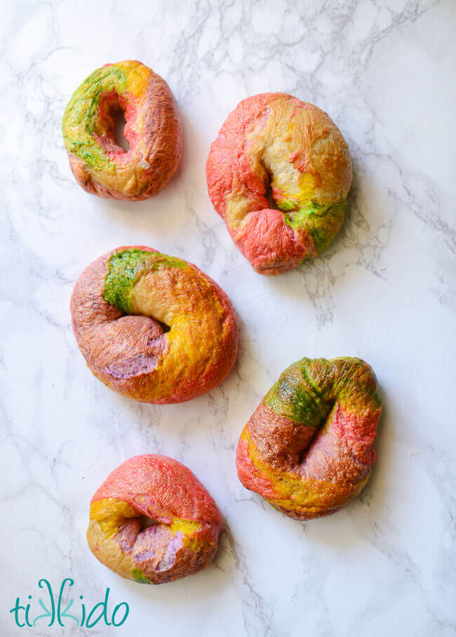 Five rainbow bagels on a white marble background.