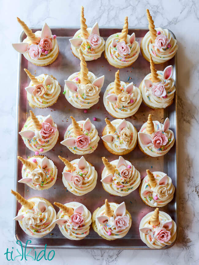 A metal tray with 20 unicorn cupcakes on a white marble surface.