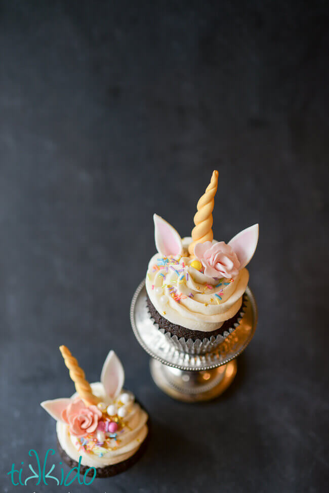 Two unicorn cupcakes decorated with gum paste unicorn horns, ears, flowers, and colorful sprinkles on a black chalkboard background.