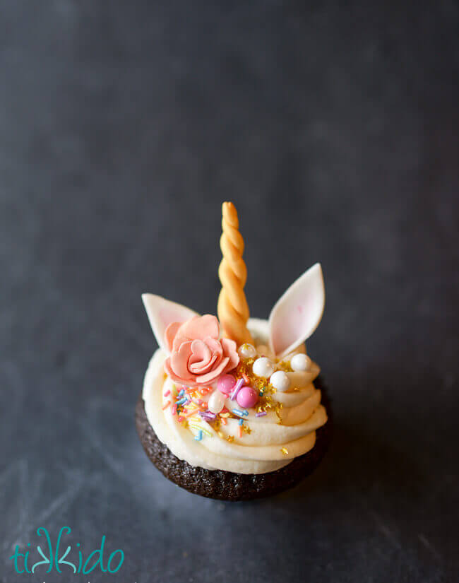 One chocolate cupcake topped with white icing, colorful sprinkles, and a gum paste rose, unicorn ears, and unicorn horn.
