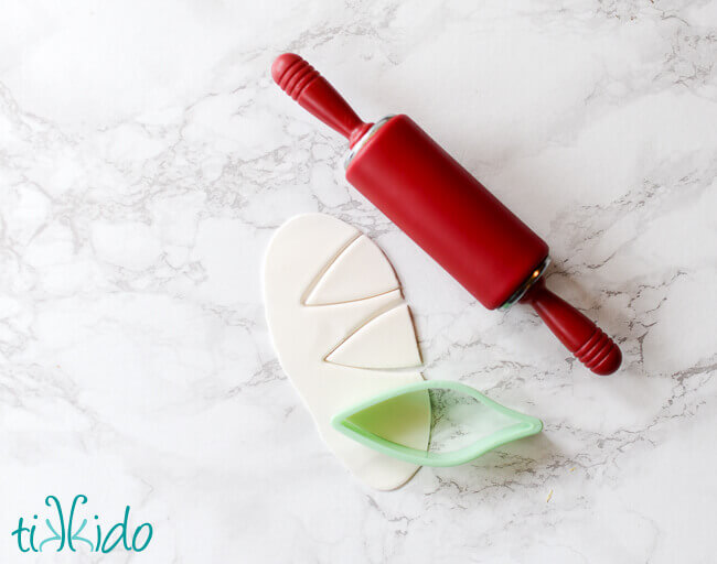 Small red silicone rolling pin next to rolled out white gum paste being cut into unicorn ear shapes with a leaf shaped gum paste cutter.