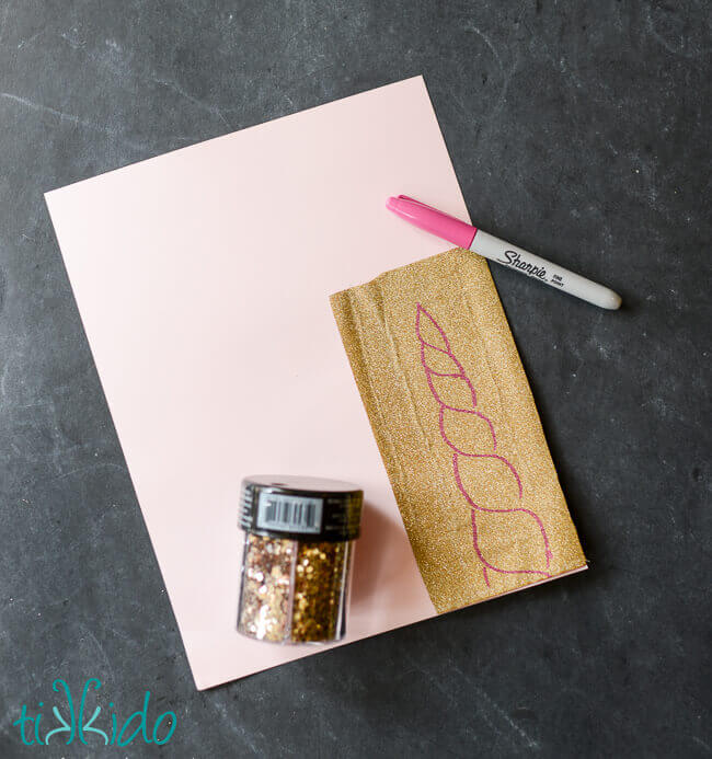 Gold paper layered on cardstock, with a unicorn horn sketched on the gold paper with a sharpie marker, and a container of glitter beside it.