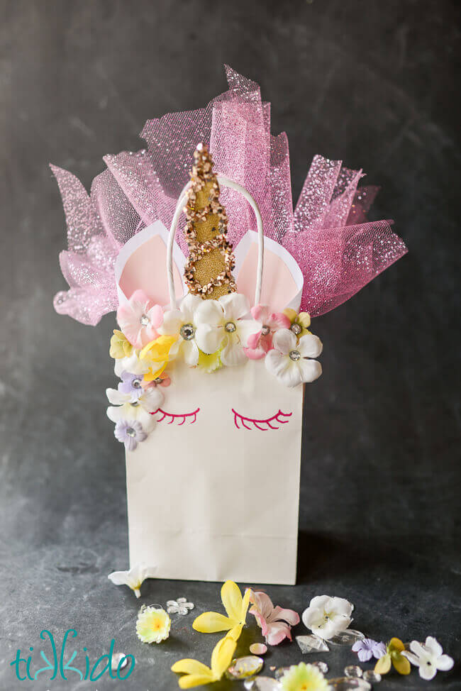 Unicorn party favor bag made out of a plain white paper gift bag, with a flower and rhinestone mane, glittery horn, paper ears, and sparkling tulle sticking out of the bag.