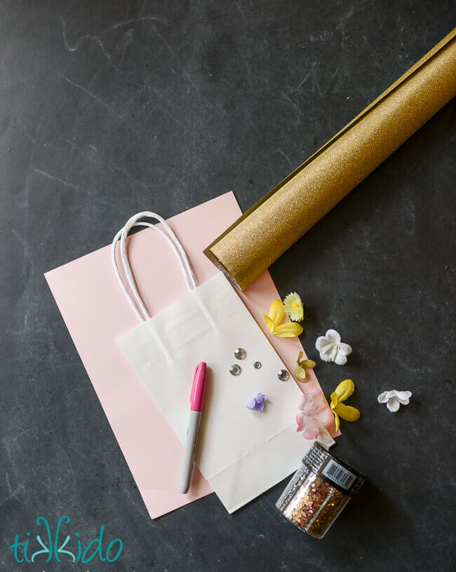 Overhead view of materials for Unicorn gift bag, including plain white gift bag, pink and white paper, golden paper, silk flowers, glitter, rhinestones, and a sharpie marker.