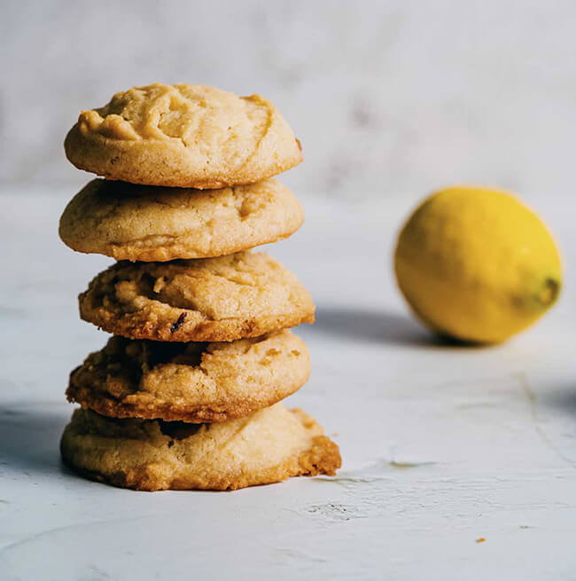 Stack of five soft vegan lemon cookies on a grey surface, with a whole lemon in the background.