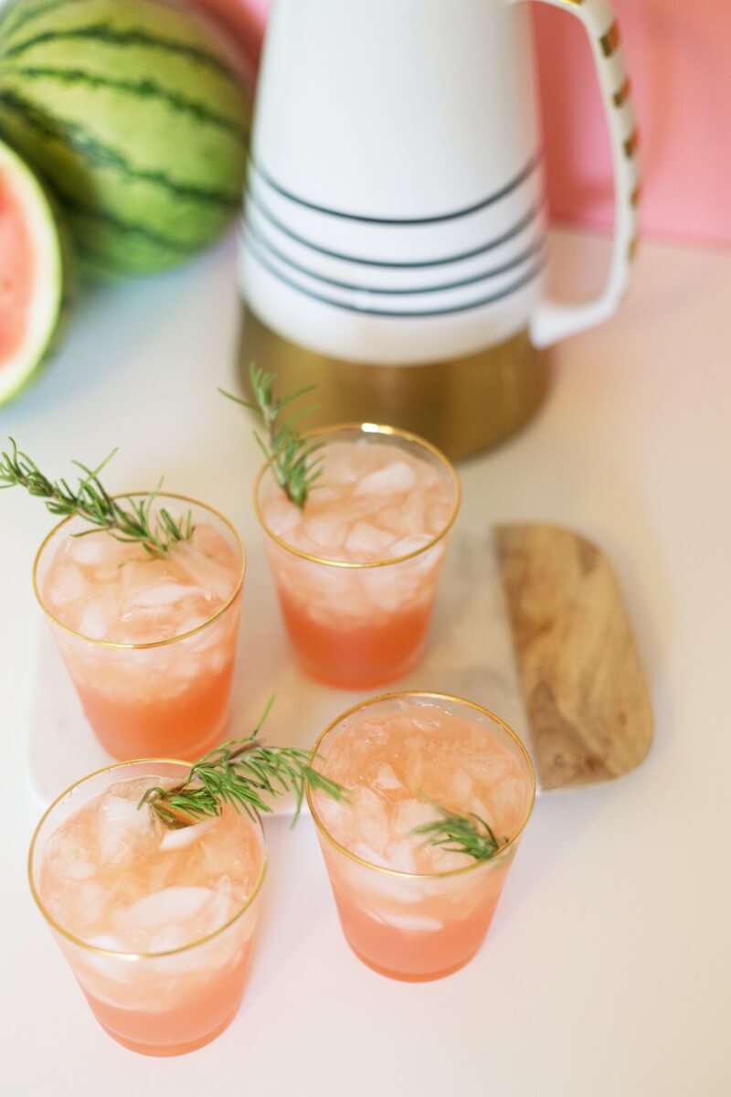 Four glasses of a blush pink cocktail garnished with a sprig of fresh rosemary.  A pitcher of the spiked watermelon rosemary punch and a sliced watermelon sit behind the glasses.