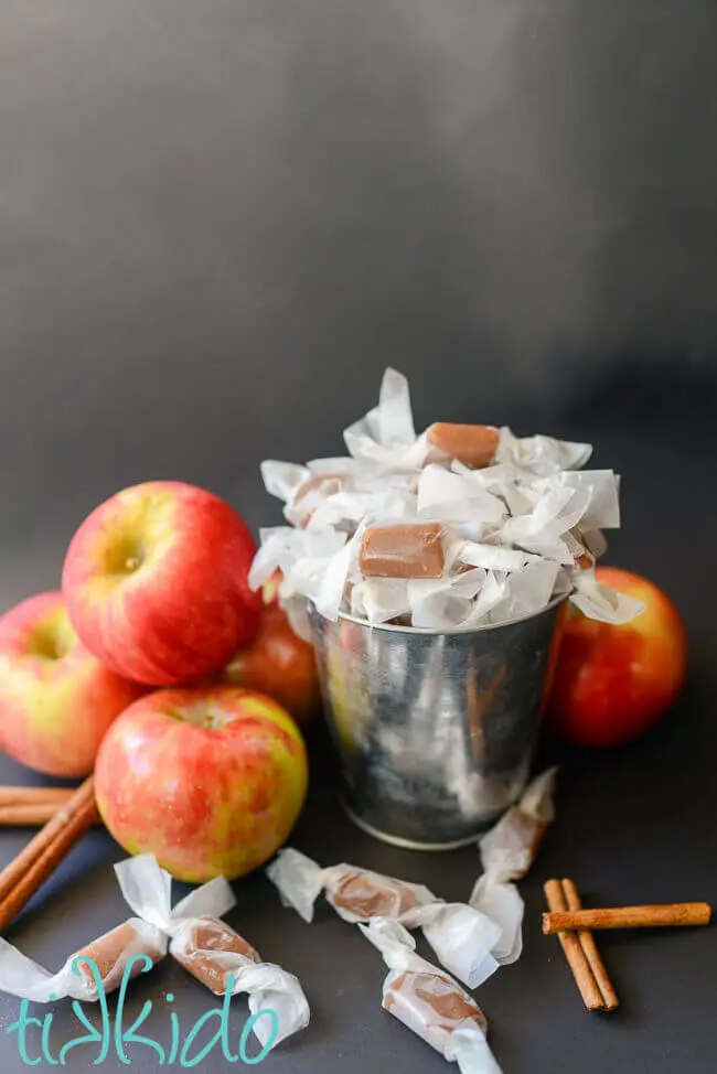 Apple cider caramels in a silver bucket, surrounded by fresh apples and cinnamon sticks.