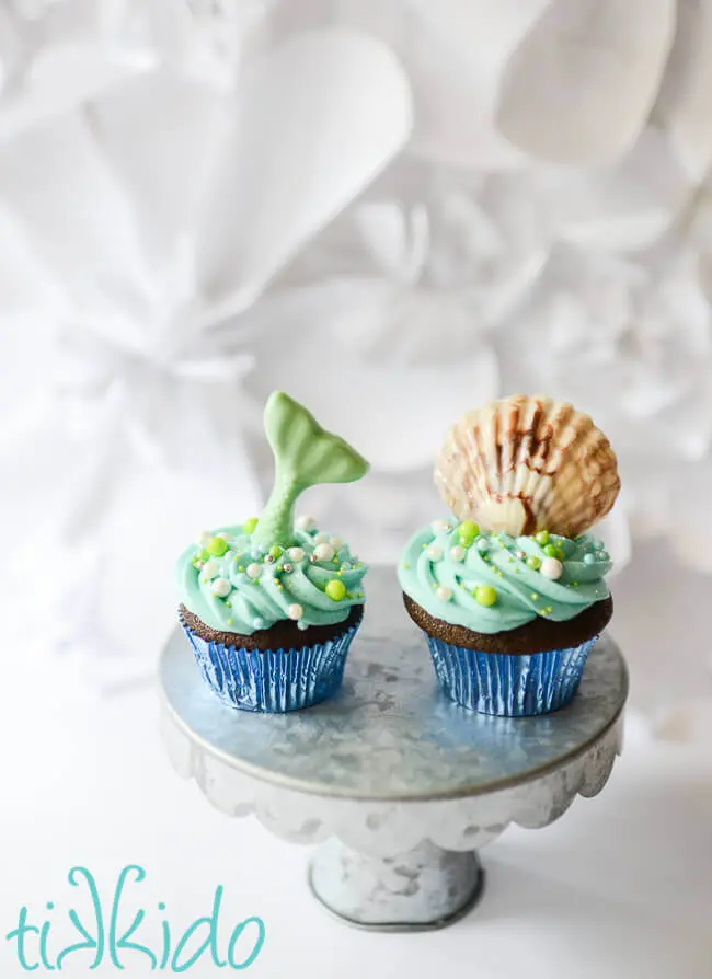 Two cupcakes, topped with a chocolate mermaid tail and chocolate shell, American buttercream icing, and sprinkles.