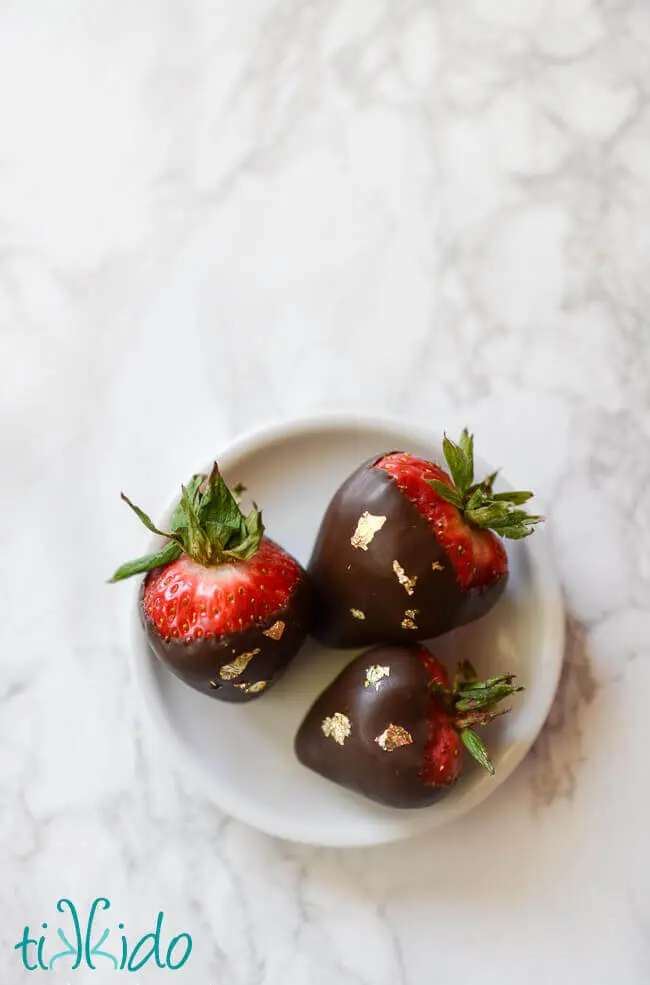 Chocolate covered strawberries decorated with edible gold leaf on a small white plate.