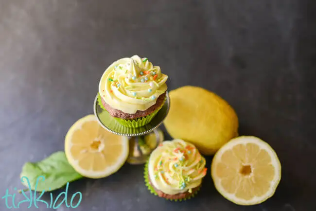 Two cupcakes topped with Lemon Buttercream Frosting and sprinkles, surrounded by a whole lemon and a lemon cut in half.