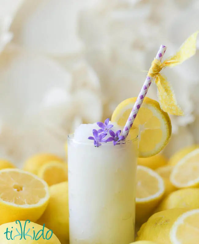 Glass of frozen lemonade with a purple straw, garnished with a slice of lemon and three purple edible flowers, surrounded by whole and halved lemons.