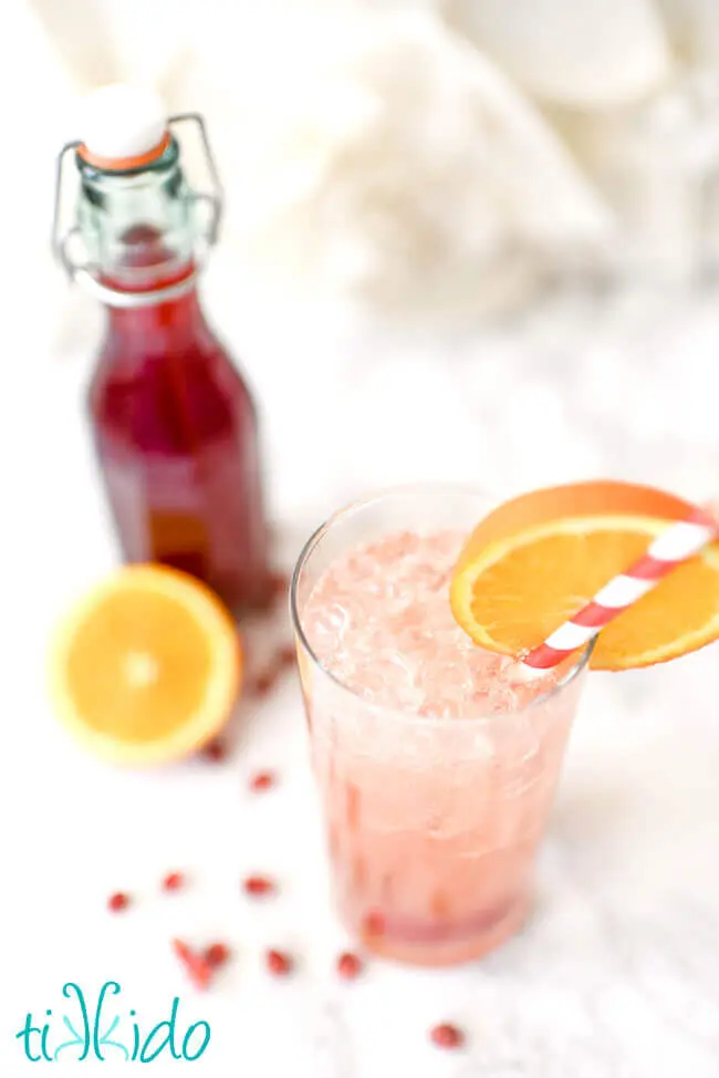 Blush pink shirley temple black cocktail in a tall glass, garnished with an orange slice and pink and white striped paper straw.  A bottle of homemade grenadine syrup, sliced orange, and fresh pomegranate seeds are in the background.