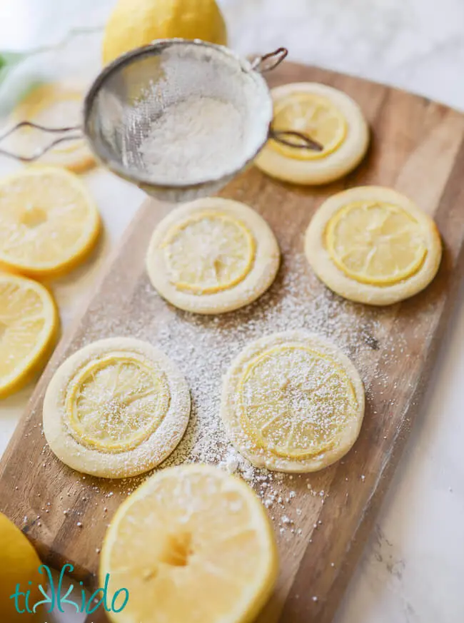 Sugar cookies  with thin slices of lemon embedded in them on a wooden cutting board, being dusted with powdered sugar from a seive.