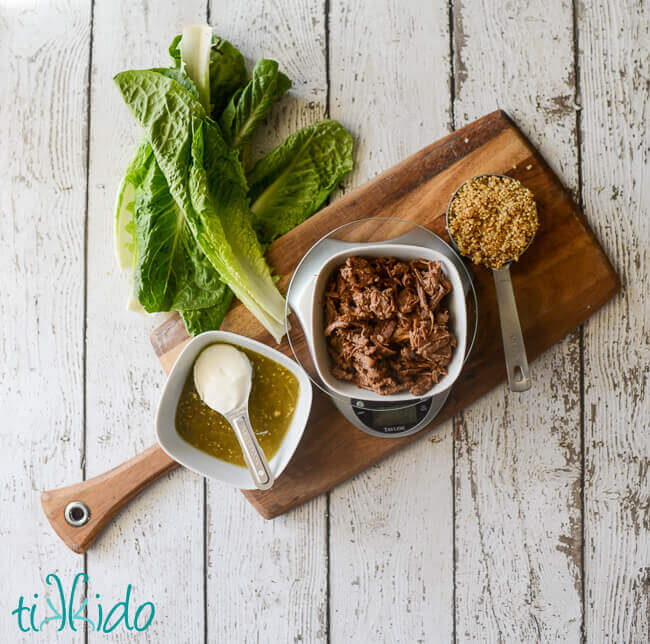 Shredded beef on a kitchen scale, cooked quinoa in a half cup measure, green salsa in a bowl, sour cream in a Tablespoon, three leaves of romaine lettuce, on a cutting board.