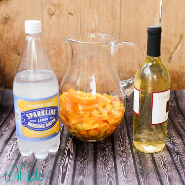 Fruit for White Sangria Recipe in a pitcher, next to a bottle of white wine and sparkling mineral water.
