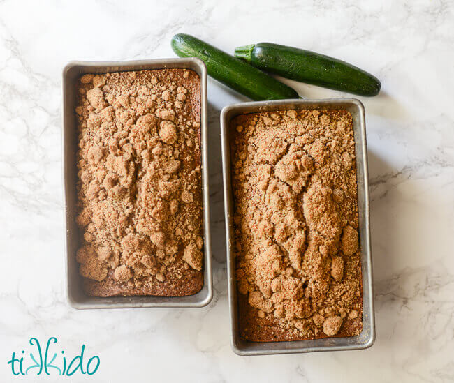 Two loaves of baked zucchini bread on a white marble surface next to two whole zucchinis.