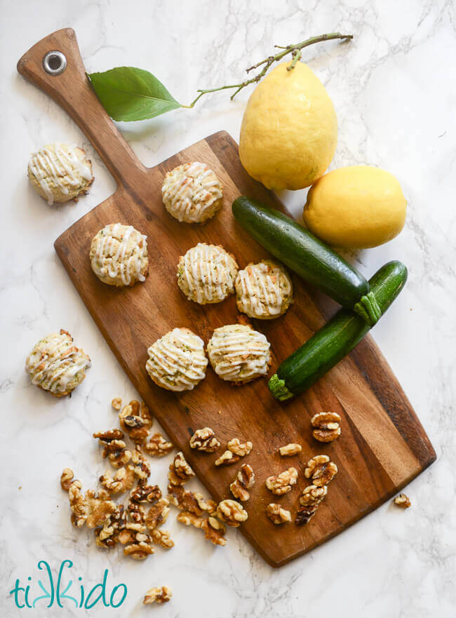 Zucchini cookies with lemon glaze on a wooden cutting board, surrounded by lemons, zucchini, and walnuts.
