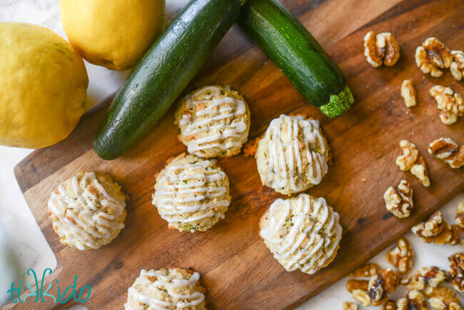 Zucchini cookies with lemon glaze on a wooden cutting board, surrounded by lemons, zucchini, and walnuts.